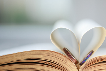 An open hardback book is pictured with two pages folded together to make a heart shape. Within the heart two wedding rings can be seen settled on the pages.
